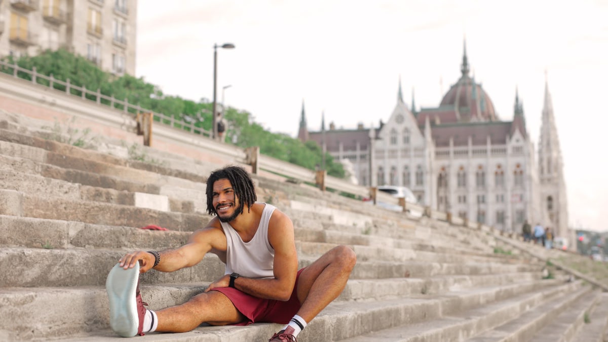 Man with dreadlocks smiling while stretching on city steps near architectural landmark.